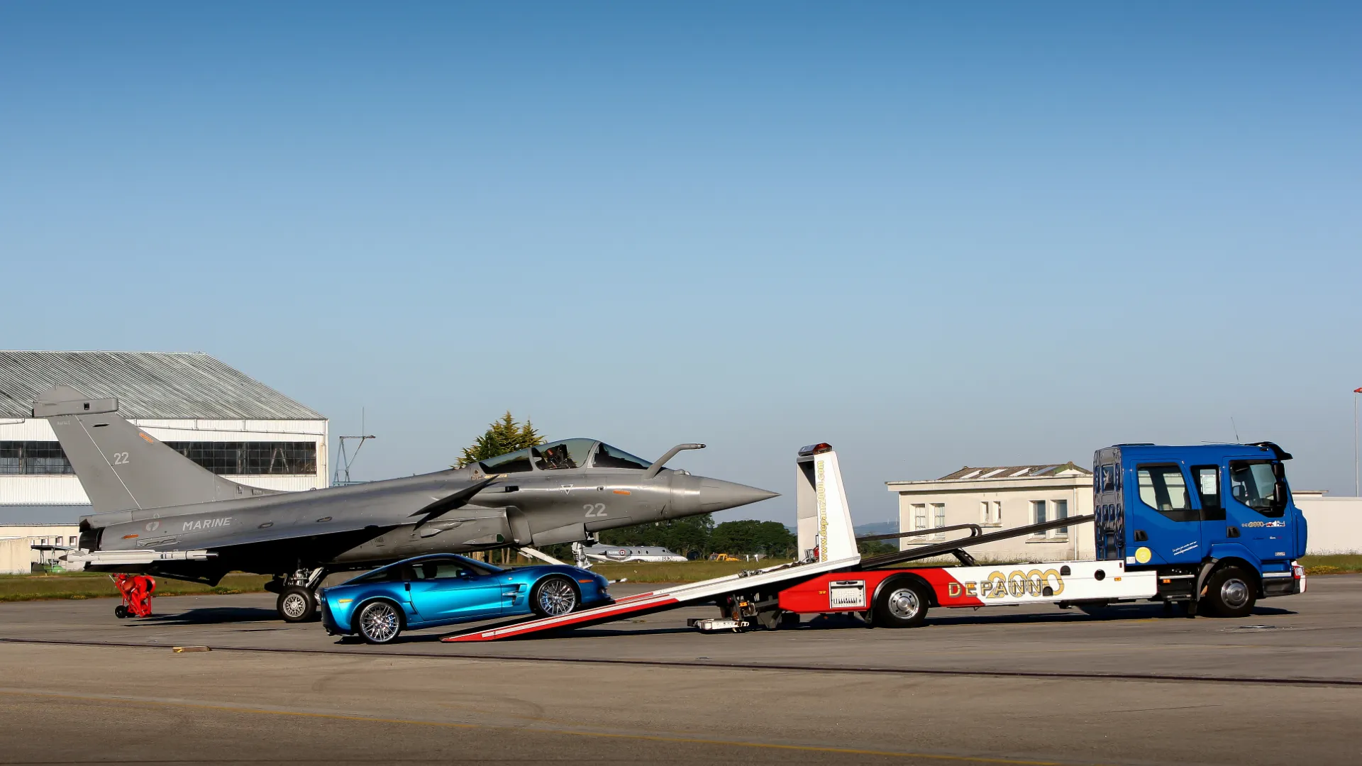 savoir-faire-histoire Camion plateau Depann 2000 chargeant une voiture de sport bleue sur un aérodrome à côté d’un avion de chasse.